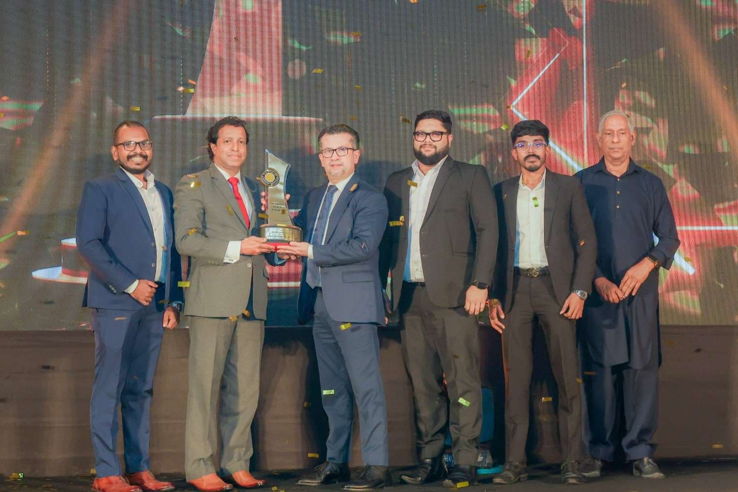 Six men in suits on a stage pose with a trophy as confetti falls during an awards ceremony.