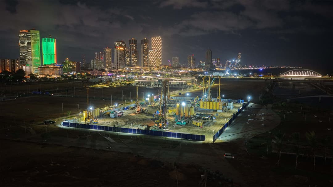 Nighttime city skyline with illuminated skyscrapers and a construction site in the foreground.